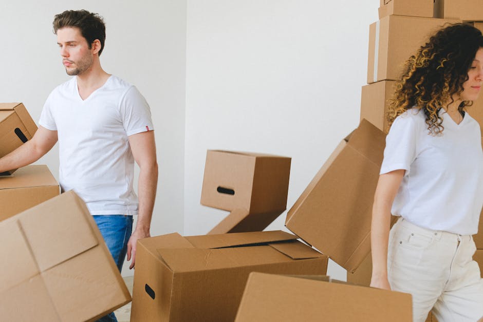 An individual engaged in a home relocation process, bending over to retrieve or place a cardboard box labeled 'pilows' in a room with natural light from large windows covered partially by dark grey cu