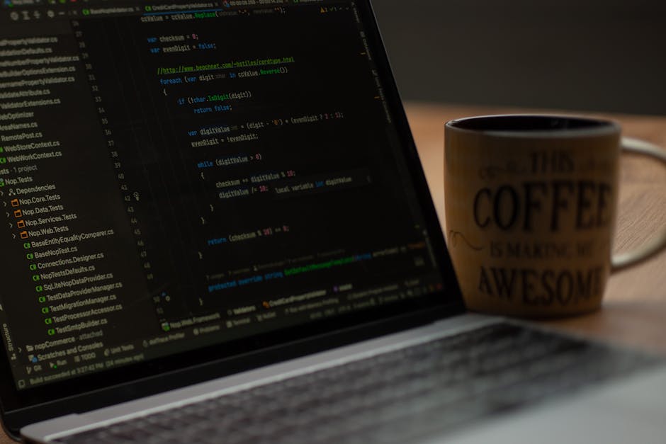 A person’s hands typing on a MacBook laptop placed on a black surface, with the screen displaying lines of code in a dark-themed integrated development environment. The background is blurred, focusi