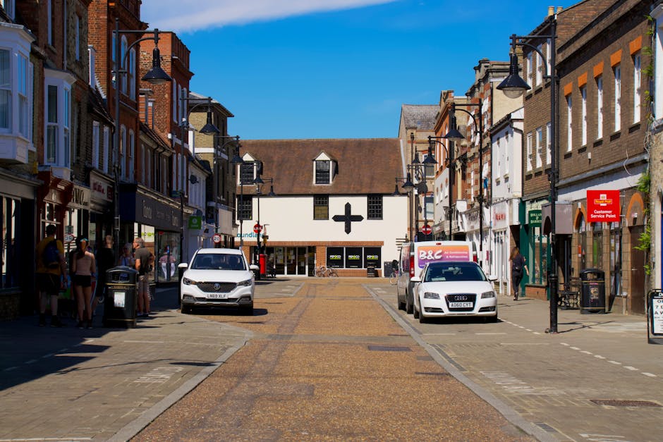 Aerial view of Uxbridge town centre showing a mix of residential and commercial buildings along High Street, with narrow streets and a nearby waterway. The scene includes parked cars, moving vehicles 